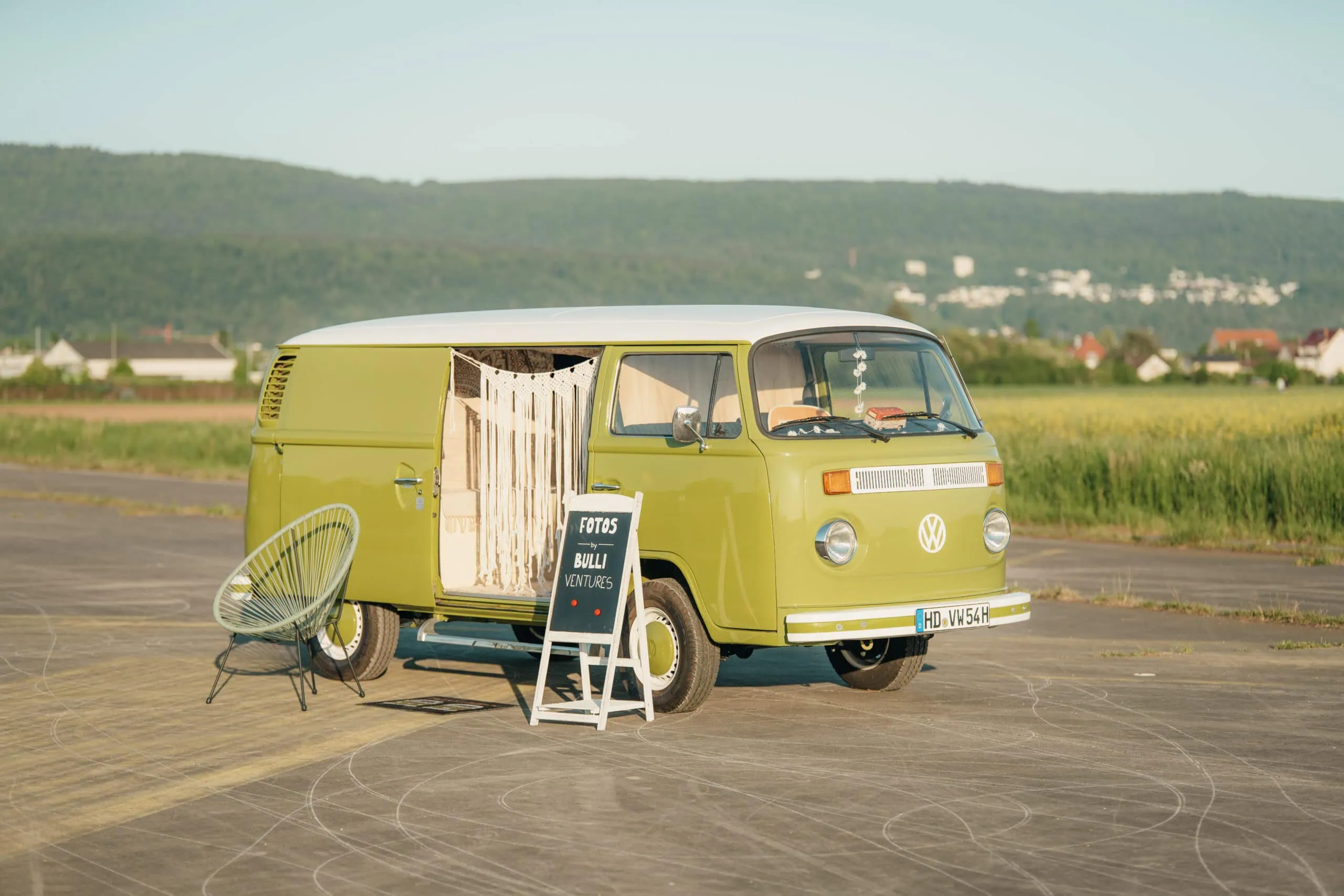 Ein grüner Volkswagen Oldtimer-Bus steht auf dem Bürgersteig, daneben ein Gartenstuhl und ein Schild.