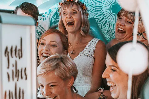 Eine Gruppe von Frauen lächelt und lacht gemeinsam in einer Fotokabine, die mit gemusterten Stoffen und Lichterketten dekoriert ist.