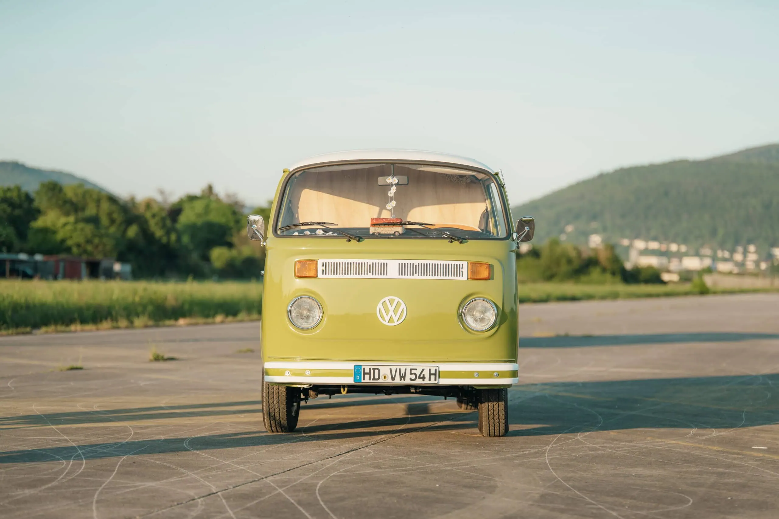 Ein grün-weißer Volkswagen-Van im Oldtimerstil steht auf einer gepflasterten Fläche, im Hintergrund Gras und Hügel.