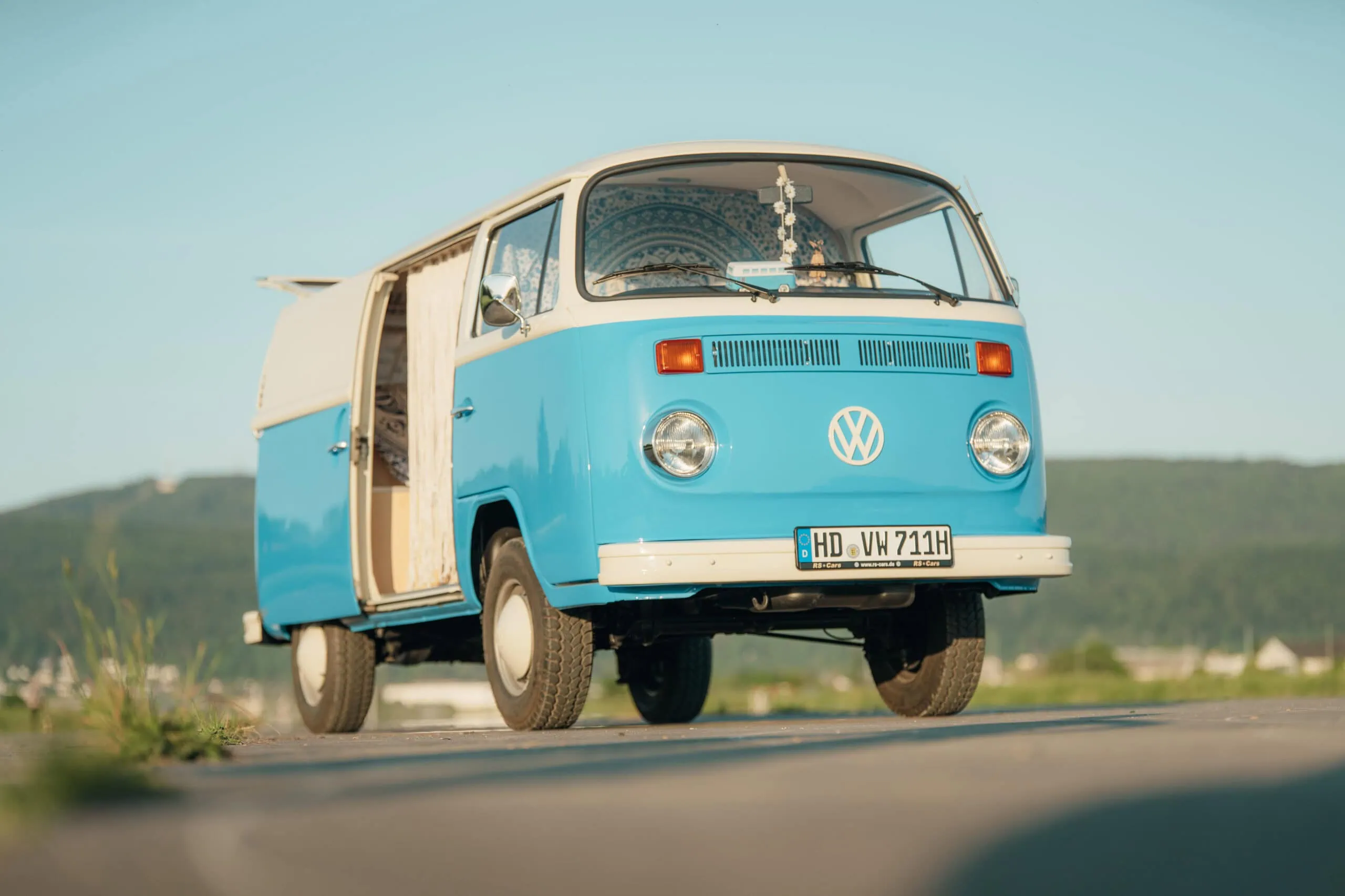 An einem sonnigen Tag steht im Freien ein blau-weißer Volkswagen-Oldtimer-Bus mit offener Seitentür.
