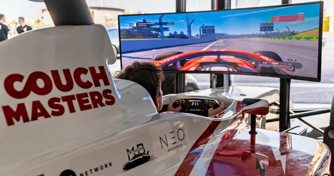 Person im Couch-Masters-Rennsimulator mit Lenkrad und Pedalen vor einem großen Bildschirm.